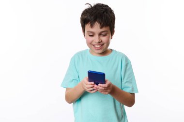 Neşeli, turkuaz tişört giyen, akıllı telefonuyla sanal oyun oynayan mutlu bir gencin beyaz arka planında izole edilmiş portresi. Dijital cihazlar ve çocuklar için eğlence aletleri. Boşluğu kopyala