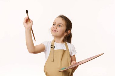 Anaokulu öğrencisinin beyaz arka planında izole edilmiş bir portre, elinde boya paleti olan sevimli bir bebek, kameranın önünde ahşap bir fırçayla resim yapıyor. Yaratıcı sanat gelişimi ve çocuk eğlencesi