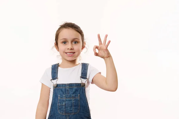 愛らしい小学生の年齢 就学前の小さな子供の女の子は 青いデニム全体で ジェスチャー 平和の兆候を示し かなり明るい歯の笑顔笑みを浮かべて カメラを見て 隔離された白い背景 スペースのコピー — ストック写真