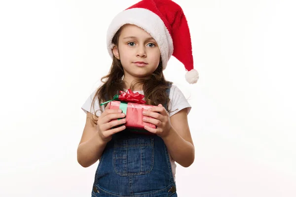 Isolated portrait on white background of a beautiful little child girl wearing santa hat and denim overalls, holding her Christmas present. Looking forward to the upcoming winter holidays. Copy space