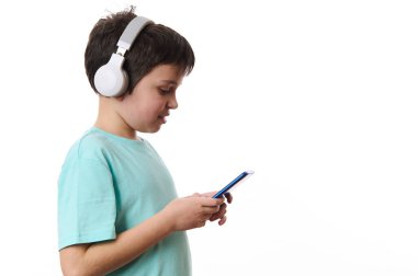 Kablosuz kulaklık takan, cep telefonu kullanan, sosyal medya içeriğini kontrol eden, internette gezinen, web sitelerine giren, beyaz arka planda izole edilmiş genç bir çocuğun yan portresi..