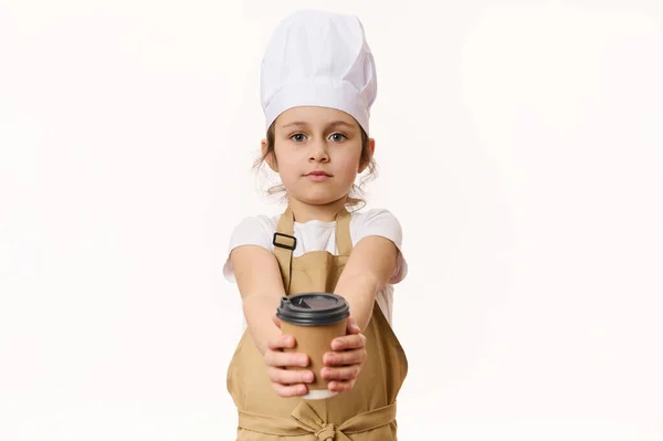 Isolated portrait on white background of cute baby girl wearing a white chefs hat and beige apron, holding, selling a hot drink in a paper cup to go, looking at camera, isolated over white background