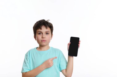 Mavi tişörtlü çocuk reklam ya da mobil uygulama için boş ekranlı akıllı telefonu işaret ediyor. Çocuklar ve dijital cihazlar. Beyaz arka planda cep telefonu olan bir gencin stüdyo fotoğrafı.