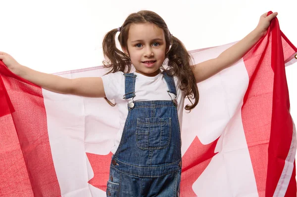 Happy little girl with two ponytails, wearing blue denim overalls, carries Canadian flag. Concept of Independence Day of Canada, the first July. Immigration. Emigration. Travel and tourism concept