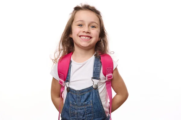 一个白种人漂亮的小学生的白色背景 一个身穿蓝色斜纹棉布工作服 背着粉色背包的小女孩的孤零零的画像 带着可爱的微笑看着镜头 — 图库照片