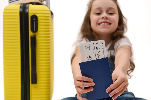 Selective focus on flight ticket or boarding pass in blurred traveler passenger childs hands near a yellow suitcase over white background. Travel. Journey. Flight. Free space for advertising text