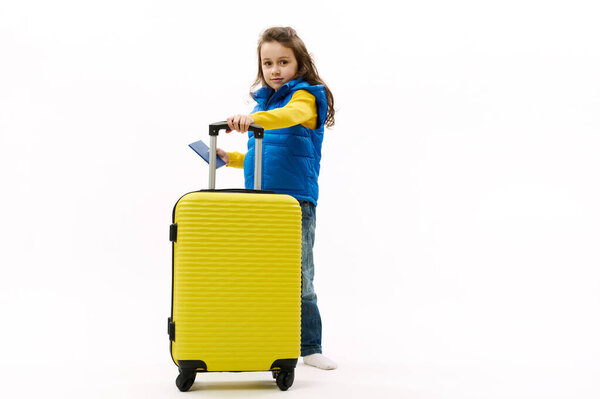 Полноразмерная маленькая девочка, в синей куртке, джинсах, держит в руках желтый чемодан и посадочный талон, изолированный на белом фоне. Туристические поездки за границу отдых отдых отдых