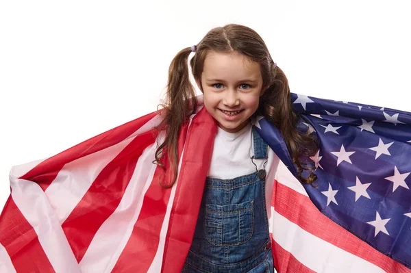 Happy citizen of USA, American little girl with two ponytails, wearing blue denim overalls, carries flag of United States of America. Concept of Independence Day. 4th July. Immigration. Emigration.