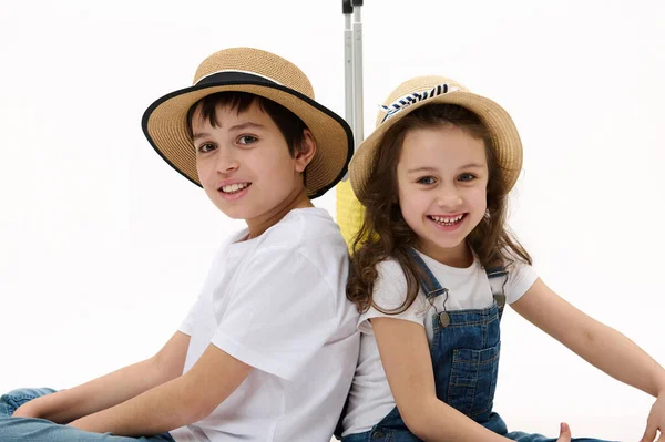 Close-up portrait on white background of lovely kids, teenage boy and his little sister baby girl sit with their backs to each other, cutely smile on camera. Children travel abroad for weekend getaway