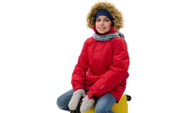 Isolated portrait on white background handsome child boy in warm red parka and blue denim jeans, sitting on a suitcase, smiling to camera. Free ad space. Winter travel. Journey. Rest getaway concept