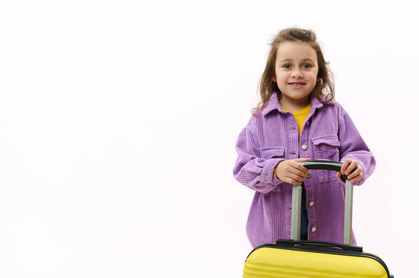 Delightful Caucasian child, lovely baby girl in violet shirt with yellow suitcase, smiling a beautiful toothy smile looking at camera, isolated on white background. Copy space. Journey Travel concept