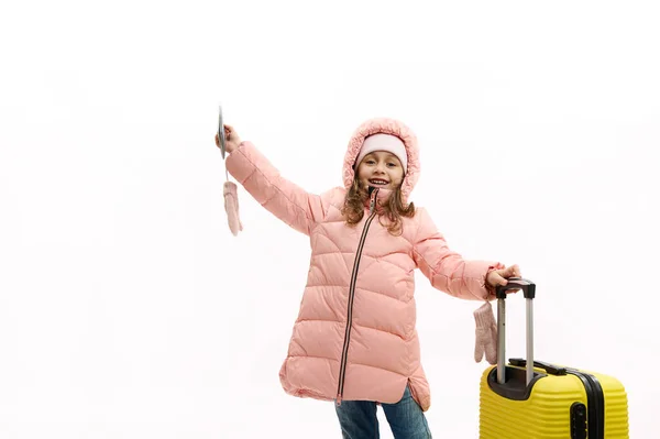 Lovely little girl in warm pink winter coat, with plane ticket, boarding pass and yellow valise, waves hello, looking at camera, isolated over white background. Weekend getaway. Travel Tourism concept