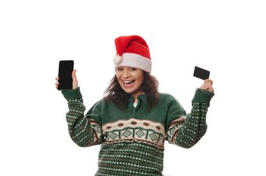 Sıcak yeşil kazaklı ve Noel Baba şapkalı, elinde siyah kredi kartı ve akıllı telefonu olan, beyaz arka planda izole bir şekilde gülümseyen neşeli bir kadın. İnternet bankacılığı, online alışveriş, mobil uygulama kavramı
