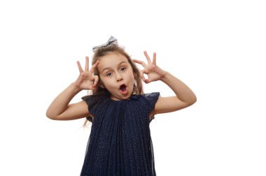 Beyaz çocuk 5-6 yaşlarında, şık giyinmiş, işaret diliyle iyi bir jest yapıyor, yüzünü ekşiterek kameraya bakıyor, beyaz arka planda izole edilmiş. Çocuk parmakları yapıyor.