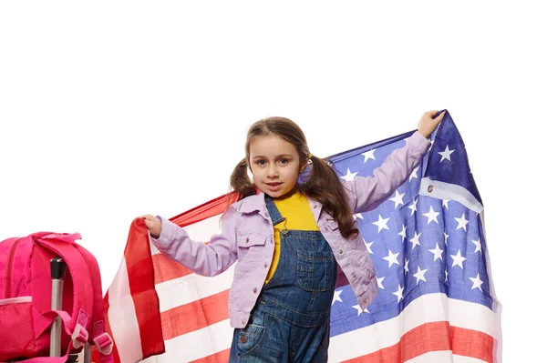 Happy little American citizen, cute baby girl in blue denim overalls, with USA flag, celebrates Independence Day on July 4, isolated on white background. Immigration, citizenship and freedom concept