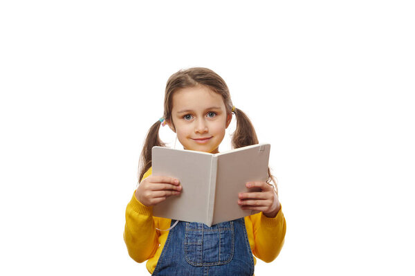 Smart first grade schoolgirl, with two ponytails, wearing a yellow pull and denim overalls, reading book over white background. Knowledge and Erudition. Back to school on new academic year semester