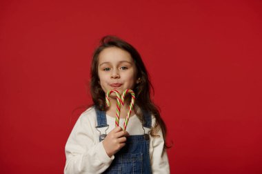 Güzel küçük kız elinde tatlı kırmızı çizgili yeşil şeker kamışı tutuyor ve kameraya tatlı tatlı gülümsüyor, kırmızı arka planda izole bir şekilde fotokopi çekiliyor. Noel ve Yeni Yıl kavramı