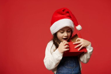 Noel Baba şapkalı 5 yaşındaki tatlı beyaz kız. Kırmızı arka planda Noel hediyesini açarken sürpriz ve olumlu duygularını dile getiriyor. Boks Günü. Mutlu yıllar konsepti