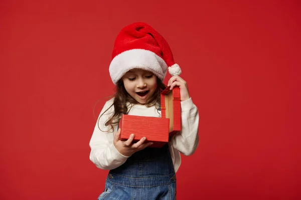 Isolated emotional portrait on red background of adorable kid, cute little girl in Santa hat, with wow emotion, expressing happiness and surprise while unpacks happy Christmas present. Copy ad space