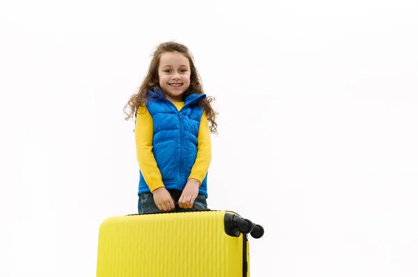 Isolated portrait on white background of a happy baby girl smiling a cheerful toothy smile, carrying a yellow suitcase, going for weekend getaway or traveling abroad to warm countries. Travel concept