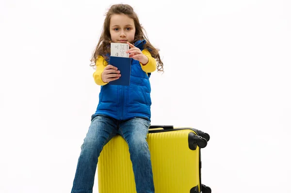 Isolated portrait on white background of a Caucasian charming kid girl in blue jacket, holding out an air ticket and boarding pass at camera, sitting on yellow travel bag, going for weekend getaway.