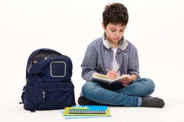 Sırt çantası ve okul malzemeleriyle beyaz arka planda oturan, ödev yapan, telif defteri üzerine kompozisyon yazan zeki bir öğrenci. Akademik yılın yeni döneminde okula dönüyorum. Bilgi Eğitimi