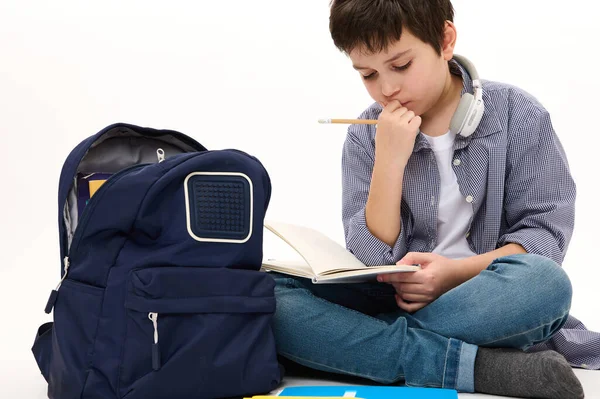 Thoughtful smart teen boy, elementary student, thinking on school project, doing homework, sitting with copybook on knees near his school bag, isolated, white background. Knowledge Education Learning