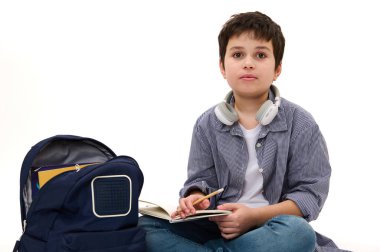 Ses kulaklığı takan, ev ödevi yapan, beyaz arka planda okul çantasının yanında oturan çok ırklı sevimli genç çocuk. Okula dönelim. Reklam alanı