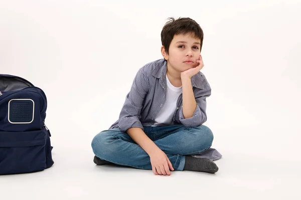 Isolated portrait on white background of a handsome smart pupil, Caucasian boy teenager in blue casual shirt and jeans, holding hand on chin, sitting near his backpack, confidently looking at camera