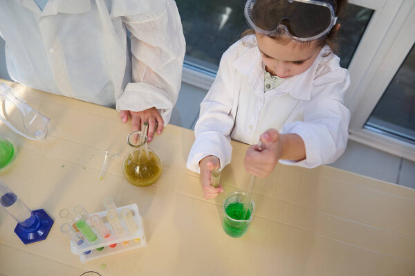 View from above of a smart school girl in white lab coat, using graduated pipette, dripping few reagents into a beaker with green chemical liquid solution, doing experiments in chemistry laboratory