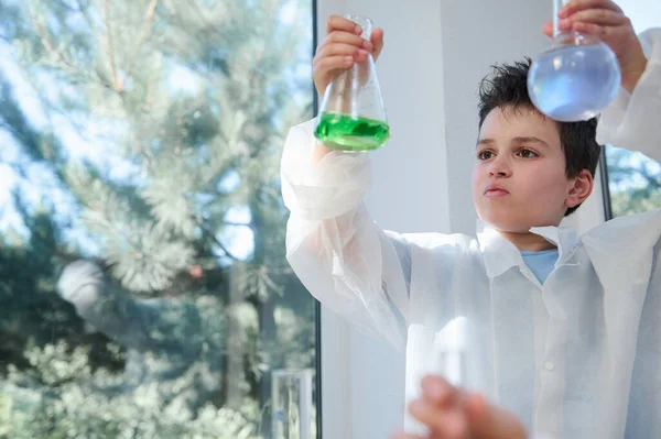 Inspired young chemist-scientist, a Caucasian smart nerd schoolboy wearing white lab coat, examining liquid chemical solutions in the lab flasks in the school chemistry laboratory. Copy space for ads