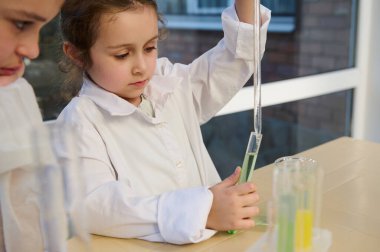 Zeki beyaz tenli, 5-6 yaşlarında küçük bir çocuk, beyaz labirent elbisesi giyen bir öğrenci, kimyasal sıvı çözeltisi ile test tüpüne az miktarda reaktör damlatıyor.