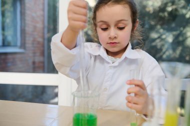 Little girl, first grade student at chemistry class, using graduated pipette and test tube, drips few reagents from beaker into test tube, conducts chemical experiments, in school chemistry laboratory