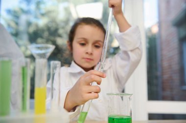 Selective focus on graduated pipette with several drops of reagent, in hands of elementary school student drips a liquid chemical into test tube, conducts experiment in a school chemistry laboratory