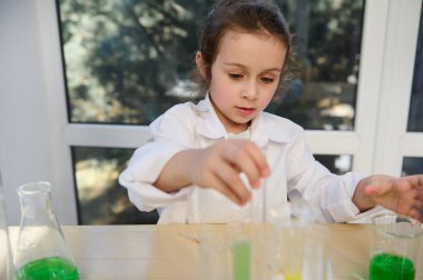 Close-up portrait of a Caucasian smart cute little girl, primary school student in white lab coat, focused on chemical experiment in school chemistry laboratory. Education. Learning. Kids development