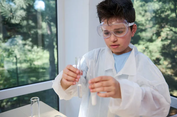 Portrait of a schoolboy, chemist scientist in goggles and white lab coat, holding test tubes with chemicals and reagents in the school chemistry laboratory. Back to school. Kids and science concept