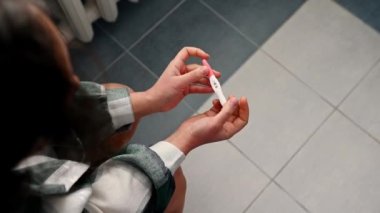 Overhead close-up view of the hands of a woman, sitting on the toilet bowl, holding a jet pregnancy test in her hands, showing a positive result. Finally pregnant. Pregnancy test in action