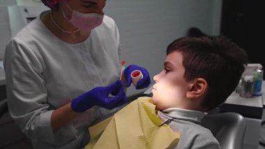 Female dentist doctor in white medical uniform and protective gloves, applying anesthetic cream to teenage boys gum before removing molar baby tooth, in modern dental clinic. Pediatric Dentistry