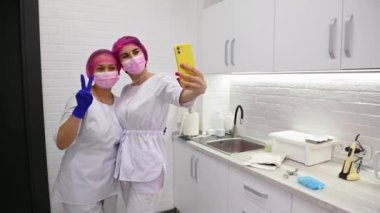 Friendly cheerful multi-ethnic team of a dentist hygienist and assistant in white medical uniform and protective mask, smiling while making selfie on smartphone in white a modern dentistry clinic