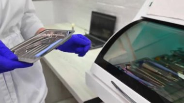 Dentist assistant in blue surgical gloves, puts a metal tray with stainless steel dental tools inside UV autoclave sterilizer. Ultraviolet thermostat for sterilizing dentistry and surgery instruments
