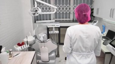 Rear view of a dentist doctor walking through the dentists office to the dental equipment, sitting down in a chair and smiling at camera, welcoming the patient for dental check up in dentistry clinic