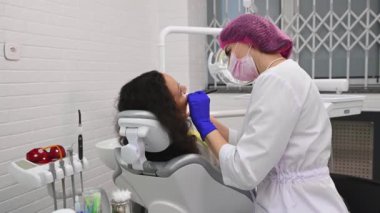 Side view female doctor dentist hygienist, using dental tools, checks the teeth of a woman patient sitting on dentists chair, in dental examination room of a modern white interior of dentistry clinic