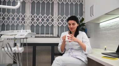 Beautiful confident Caucasian woman, dentist orthodontist doctor relaxing after hard working day in dentistry clinic, sitting on the chair and enjoying her hot aroma coffee in the dentists office