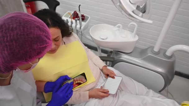 医生正牙医生向坐在牙医上的病人解释安装假牙 瓷窗的过程 并在现代智能手机上展示一些照片 牙科整形外科 — 图库视频影像