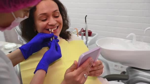 在牙医座位上 密切观察微笑的女性病人 医生正牙时看着小镜子 根据颜色图选择牙龈的阴影 美容美发牙医 牙科治疗 — 图库视频影像