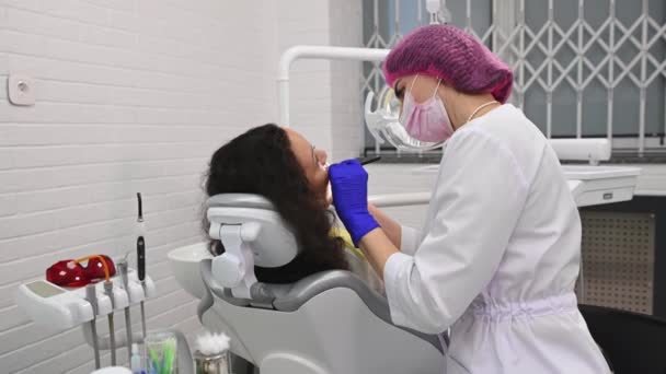 在牙科诊所的一个现代化的白色内部牙科诊所的牙科检查室里 女医生卫生学家用牙科工具检查坐在牙医椅子上的女病人的牙齿 — 图库视频影像
