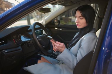 Confident stylish Caucasian middle-aged pretty woman, checking her modern smartphone, while sitting in the car on drivers seat. Business woman driving car in urban road. People Business Lifestyle