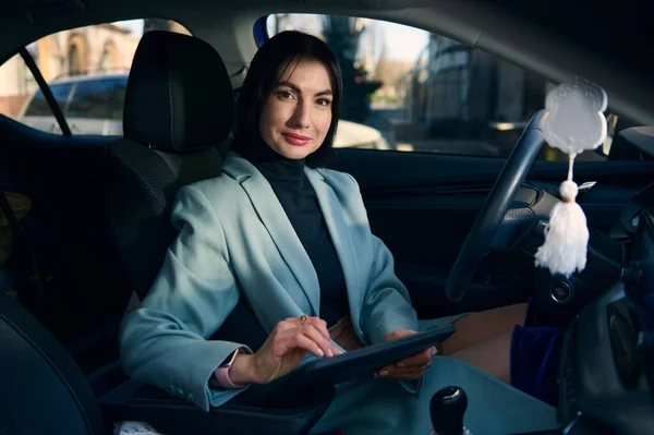 Confident portrait of a Caucasian middle-aged, successful business woman, female boss, executive director, supervisor, sales manager with a digital tablet, looking at camera from a modern car interior