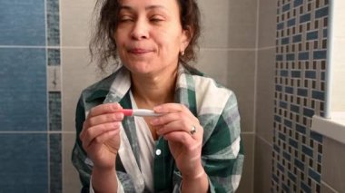 Beautiful multi-ethnic woman, sitting on toilet bowl, taking pregnancy test, anxiously awaiting the result, smiling a cheerful toothy smile, rejoicing ot positive result, showing the test on camera