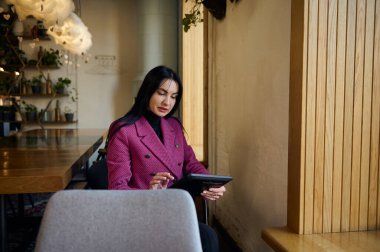 Middle-aged Caucasian successful concentrated woman boss, restaurateur, chief executive, manager using modern digital gadget, writes notes and weekly plan report in cozy interior of new restaurant
